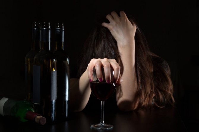 Девушка держится за голову и за бокал вина, на столе перед ней стоят пустые бутылки из-под вина
