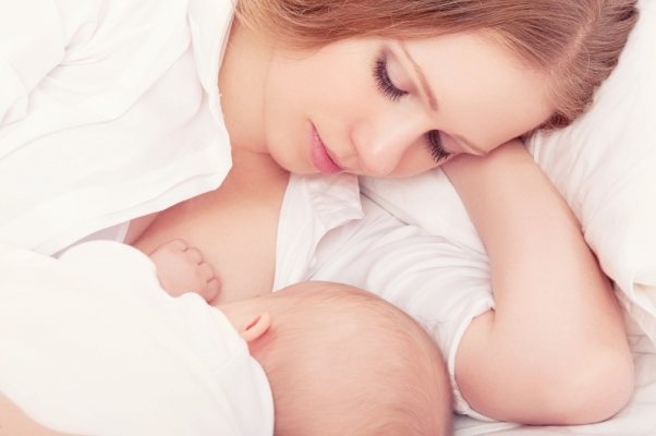 A baby in a white diaper lying down sucks his mother&#39;s breast in a white shirt