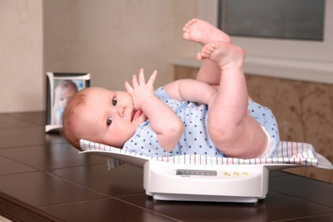 A child in a blue bodysuit sucks his finger and lies on the scales